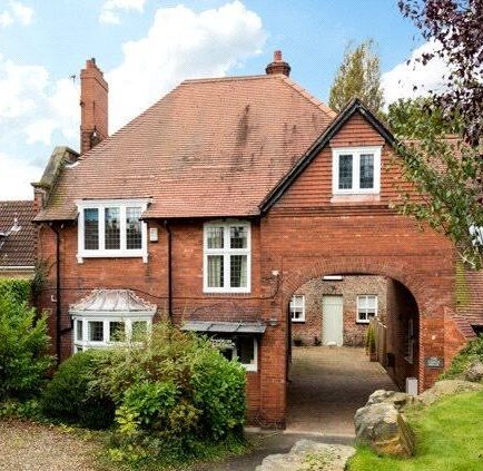 A brick house with a large arch and several tall chimneys sits at the end of a driveway.