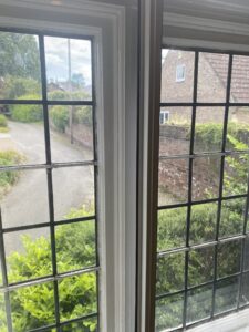 A pair of white-framed wooden windows with leaded glass panes look out over a cluster of bushes and a driveway.